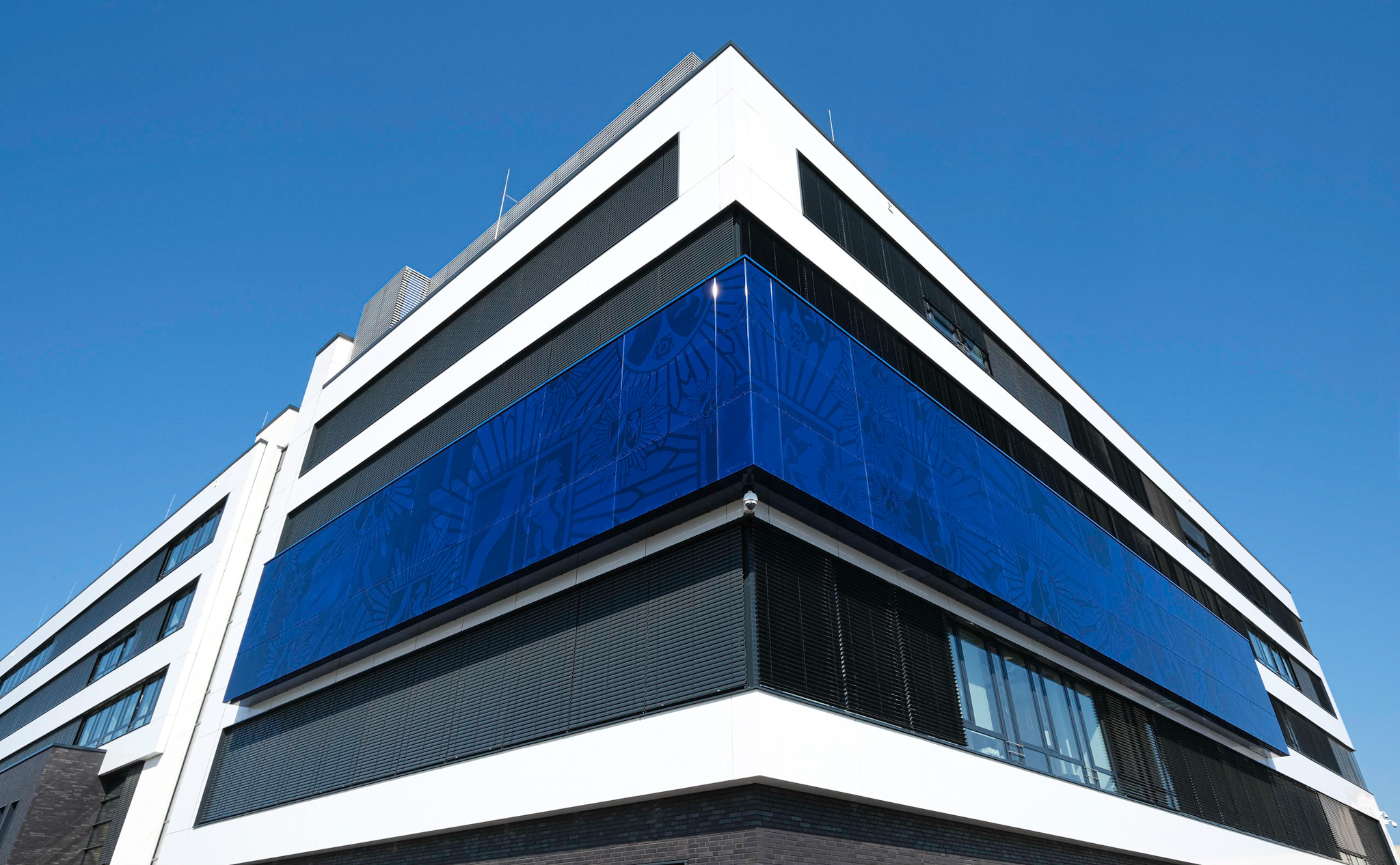 Ein weißes Gebäude mit schwarzen Fenstern und Polizeiblauer Fassadenverzierung wurde aus der Froschperspektive vor einem blauen, wolkenlosen Himmel Fotografiert. In der Fassadenverzierung lässt sich das Logo der Landesregierung NRW erkennen, welches je nach Lichteinfall sehr blass sichtbar wird. 