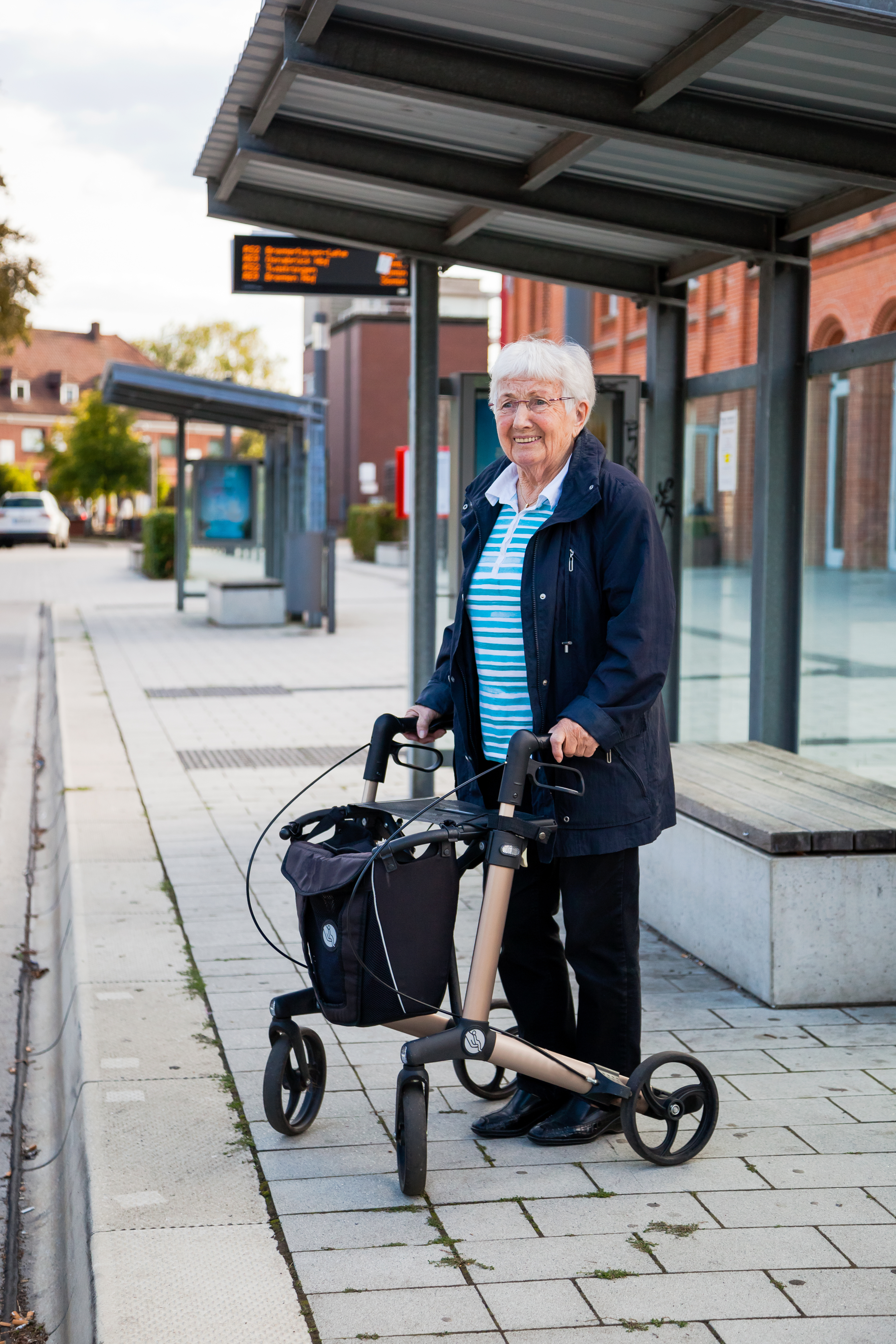 Ältere Frau steht mit ihrem Rollator an einer Bushaltestelle.