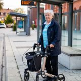 Ältere Frau steht mit ihrem Rollator an einer Bushaltestelle.