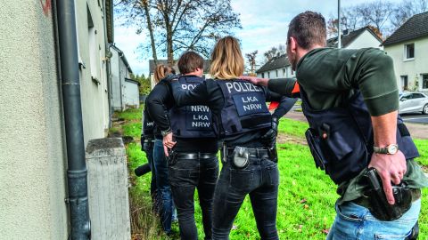 A group of five police officers are standing in a row next to the wall of a house. They each have a hand on the shoulder of the person in front of them. They are standing slightly crouched and wearing vests that say LKA NRW on them. Trees and other houses can be seen in the background.