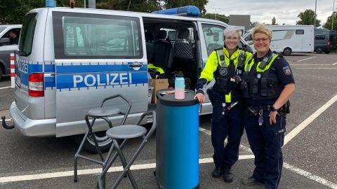 Zwei Polizistinnen in Uniform stehen an einem Stehtisch mit zwei Stühlen vor einem Streifenwagen der Polizei und lächeln in die Kamera.
