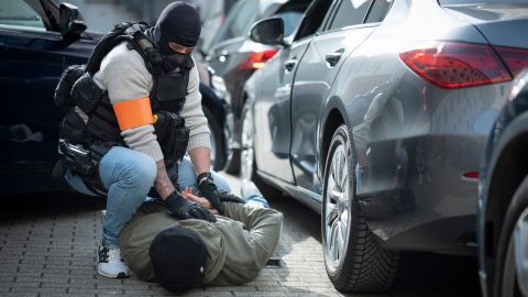The driver of the stopped car is lying on his stomach on the ground. His arms are crossed behind his back. An officer is kneeling next to him and holding his arms.