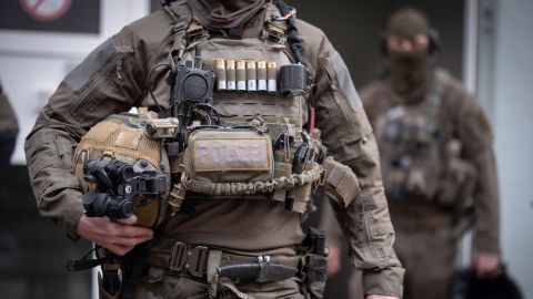 In the foreground, an SEK officer can be seen in a cut-out from his neck to his thighs. He is wearing a uniform and numerous items of equipment, as well as several weapons. Another SEK officer can be seen in the background.
