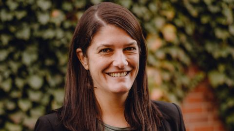 The portrait shows Anne Herr, head of the staff unit of the #Sicher Im Dienst state initiative in Münster. She is smiling. An overgrown wall can be seen in the background.