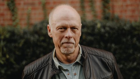 You can see a portrait of police chief inspector Ralf Hövelmann. He is wearing a shirt and leather jacket. An overgrown wall can be seen in the background.