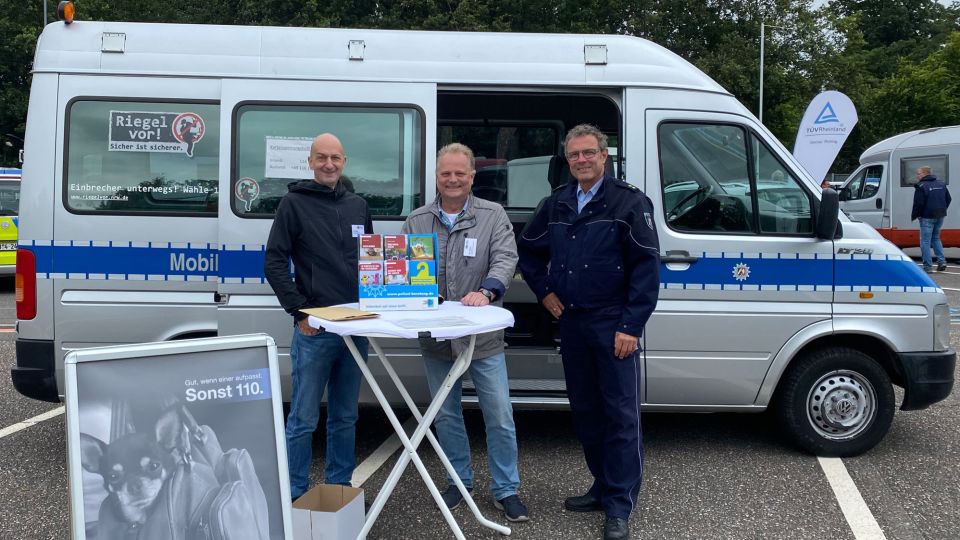 Zwei Kollegen der Kriminalprävention der Polizei Mönchengladbach stehen an einem Stehtisch vor einem Streifenwagen gemeinsam mit dem Leiter der Direktion Verkehr der Polizei.