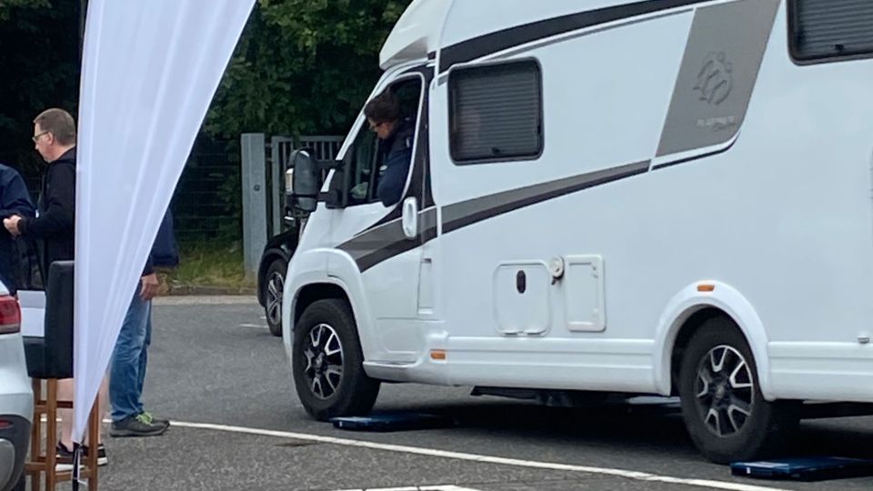 Ein weißes Wohnmobil fährt gerade von den Messplatten der Radlastwaage des TÜV Rheinland herunter. Links neben dem Wohnmobil steht ein Mitarbeiter des TÜV.