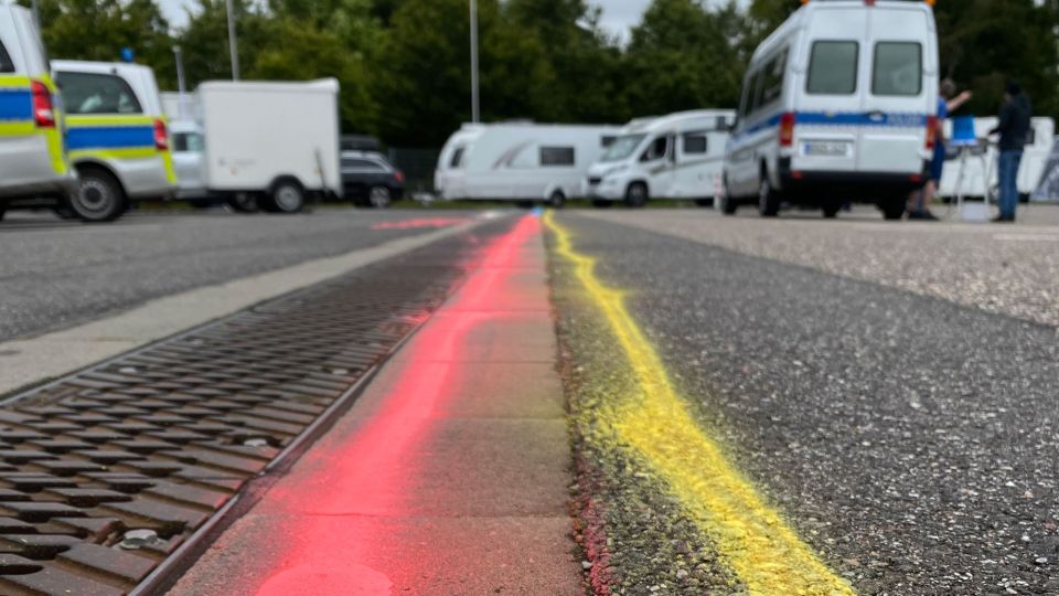 Das Foto wurde sehr nah am asphaltierten Boden aufgenommen. Auf dem Asphalt sind ein roter und ein gelber Strich zu sehen, diese laufen in den Hintergrund. Im Hintergrund sieht man verschwommen mehrere Wohnmobile, Streifenwagen der Polizei und Menschen stehen.