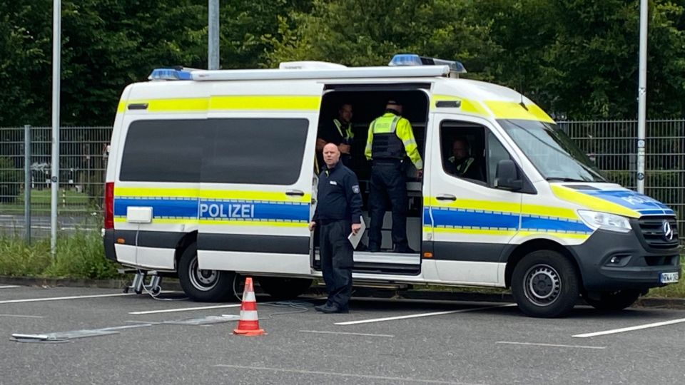 Ein großer Polizeibus mit blauer und neon-gelber Folierung steht mit geöffneter Seitentüre auf einem Parkplatz. Auf dem Boden liegt eine Radlastwaage und zwei Polizisten stehen an dem Fahrzeug.