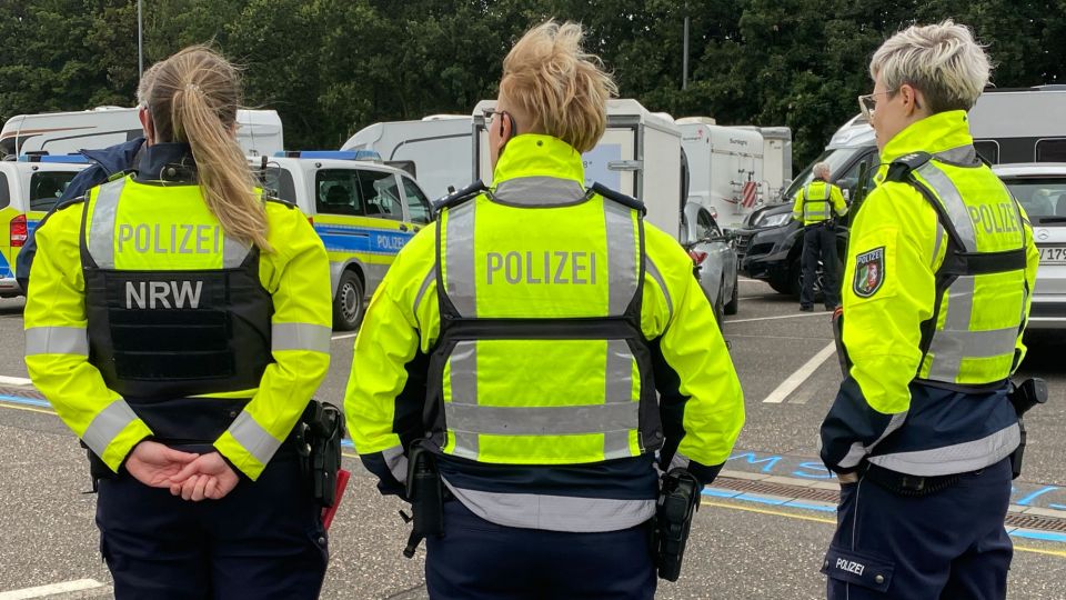 Drei Polizistinnen sind in Uniform von hinten zu sehen, wie sie mit einem älteren Herren sprechen und ihn zur Verkehrssicherheit beraten. 