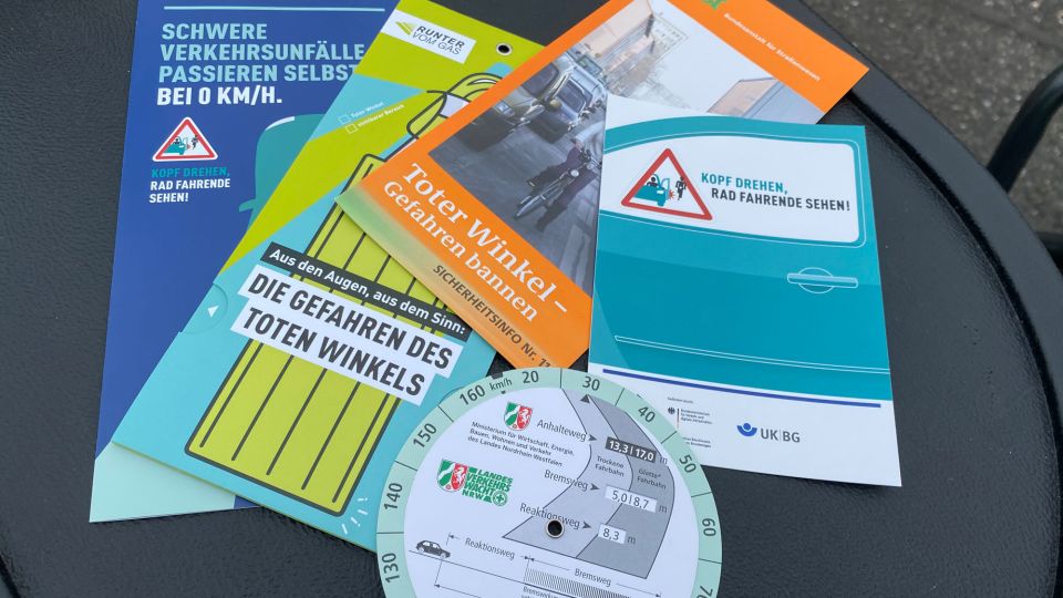 Auf einem schwarzen Untergrund liegen vier Informationsflyer überlappend. Sie behandeln die Themen Bremsweg, Toter Winkel und Fahrradfahrer im Straßenverkehr.