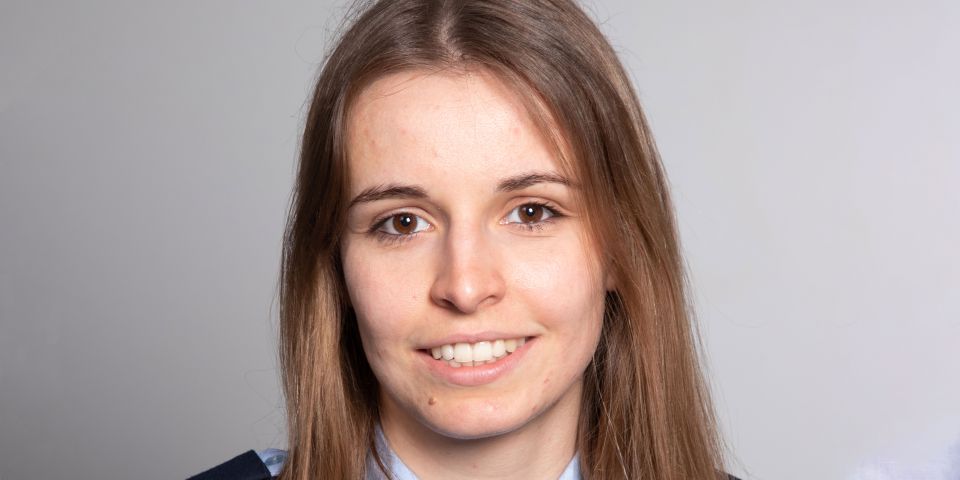 Brünette Frau in hellblauer Polizeiuniform lächelnd vor grauem Hintergrund.